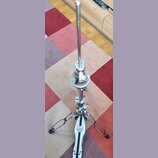 Stříbrný kovový pedál na hi-hat značky MAPEX. Nástroj stojí na vícebarevném koberci a je vybaven kovovou tyčí a mechanismem s utahovacími šrouby. Pov…