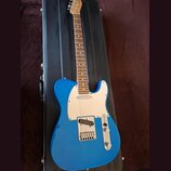 Modrá elektrická kytara typu Telecaster s bílým pickguardem a hnedým hmatníkem. Nástroj leží v černém tvrdém kufru. Na hlavě kytary je viditelné logo…