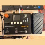Černý kytarový procesor s připojeným expresemi pedálem. Na zařízení je nápis MG-400 MODELING GUITAR PROCESSOR a oranžový štítek TS/AC HD CORE IMAGE.…