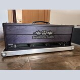 Černý transportní kufřík s dřevěným předním panelem a hliníkovým rámem. Na panelu je bílé logo s nápisem SALVATION AUDIO a grafickým symbolem vlny. V…
