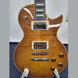 Elektrická kytara s trupem z plaménovky v žlutohnědém gradientu. Nástroj má dva humbuckery s krémovými rámečky, Tune-o-matic kobylku a stopbar struno…