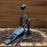 Fotografie znázorňuje jednoduchý basový pedál v černé barvě s kovovými prvky. Na stupni je bílý nápis TAMA a stylizované písmena DN, pod nimiž je men…