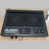 Černý elektronický samplepad s dvěma velkými gumovými pady. Na přístroji je bílý nápis ALESIS SAMPLEPAD 4 a v pravé části ovládací prvky s popisky OP…