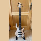 Elektrická basová kytara s přírodním dřevěným povrchem těla a hmatníkem z tmavého dřeva. Na hlavě je viditelné logo "Ibanez". Nástroj má dva humbucke…