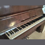 Tmavě hnědé dřevěné piano s klávesnicí. Povrch dřeva vykazuje známky opotřebení a drobné škrábance. Na klávesnici jsou vidět nečistoty a mírné změny…