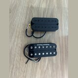 Dva elektrické kytarové snímače typu humbucker v černé barvě. Horní snímač má černé plastové krytky s viditelnými poloslnoidy. Dolní snímač má stříbr…