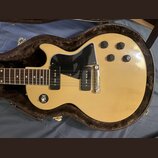 Světle žlutá elektrická kytara s černým pickguardem a čtyřmi ovládacími knoflíky v hnědém plišovém pouzdře. Nástroj má dva černávé humbuckery a stříb…