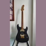 Černá elektrická kytara s matným povrchem a zlatým hardwarem. Na hlavě hmatníku je viditelné logo "Cort". Nástroj má dva humbuckery a zlatý most. Kyt…