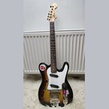 Elektrická kytara s trupem v sunburst provedení a bílým pickguardem. Na hlavě hmatníku je nápis Squier STRAT a logo Fender. Trup vykazuje výrazné opo…
