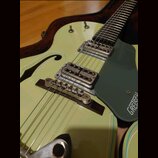 Světle zelená polodubová elektrická kytara s tmavě zeleným plechtrátorem. Na plechtrátoru je viditelný bílý nápis GRETSCH. Nástroj má dva snímače s k…
