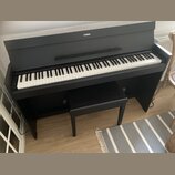 Černý digitální piano s klaviaturou a přívodním kabelem. Na horní části korpusu je bílé logo YAMAHA. Před nástrojem stojí černá pianová stolička. Pov…