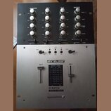 DJ mixážní pult s černým horním panelem a stříbrným spodním tělem. Na přístroji jsou bílé ovratové knofíky a přepínače s nápisy MAIN MIC, CH-1, CH-2…