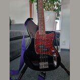 Černá elektrická basová kytara s tmavě hnědým hmatníkem. Nástroj má červený pickguard s texturou imitující dřevo a dva snímače. Na těle není vidné žá…