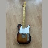 Elektrická kytara typu Telecaster s javorovým hnedým krčkem a korpusem v tmavě hnědém sunburst provedení. Na hlavě krku je viditelné logo Fender. Bíl…
