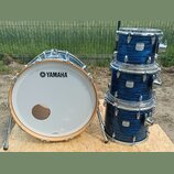 Modrá bicí souprava s kovovým hardwarem. Na bílém bendášu basového bubnu je černé logo a nápis YAMAHA. Na třech tom-tamech jsou viditelné bílé štítky…