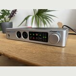 Stříbrné audio rozhraní s kovovým povrchem a velkým ovládacím knoflíkem. Na zařízení jsou nápisy ConnectAUDIO 2/4 a iConnectivity. Přední panel obsah…