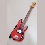 Čtyřstrunná elektrická basová kytara s červeným trupem a hnědým hmatníkem. Na korunce je viditelné logo Squier. Kytara má tmavě hnědý pickguard s tex…