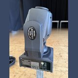 Šedý LED reflektor s pohyblivou hlavou. Na boku je černý oválný štítek s bílým nápisem MINI LEDSPOT 60W MOVING HEAD LIGHT. Spodní část obsahuje ovlád…