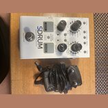 Bílý efekt pedál s nápisem SDRUM Strummable Drums. Zařízení obsahuje dva šedé tlačítka s nápisy KICK a SNARE, několik černých otočných ovladačů a řad…