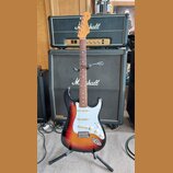 Elektrická kytara s tvarem Stratocaster, v barevné gradaci od žluté přes oranžovou po černou. Na hlavě hmatníku je viditelné logo Fender. Kytara má b…