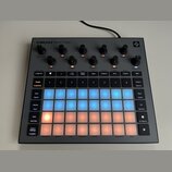 Šedý sampler s nápisem CIRCUIT RHYTHM. Zařízení obsahuje několik černých otočných ovladačů a mřížku podsvícených padů v modré a oranžové barvě. Na po…