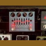 Stříbrný efekt pedál s nápisy ALPHA OMEGA ULTRA a DARKGLASS ELECTRONICS - MADE IN FINLAND. Na horní straně jsou čtyři černé ovládací knoflíky s popis…