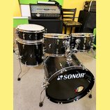 Černá bicí souprava značky SONOR. Na basovém bubně je bílé logo SONOR, na ostatních bubnech jsou viditelné štítky s nápisem ACK. V pozadí je kytarový…