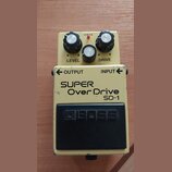 Kytarový efekt pedál v béžovém provedení s černým spínacím pedalem. Na těle jsou nápisy SUPER OverDrive SD-1, OUTPUT, INPUT a na pedálu logo BOSS. Ho…