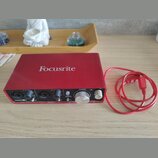 Červené audio rozhraní s nápisem Focusrite na horní straně. Na předním panelu jsou dva vstupy, ovladače zisku a knoflík s nápisy MONITOR a DIRECT MON…