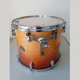 Kytarový nebo bicí buben s dřevěným korpusem v oranžovém přechodovém odstínu (sunburst). Na korpusu jsou umístěna dvě kulatá stříbrná loga s nápisem…