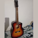 Akustická kytara s cutawayem v přechodovém žlutě-černém provedení (sunburst). Na hlavě hmatníku je viditelné bílé logo ve tvaru ptáka. Hmatník obsahu…