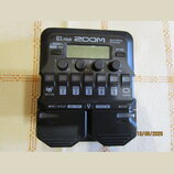 Černý multi-efektový procesor s nápisy G1 FOUR, ZOOM a Multi-Effects Processor. Zařízení má malý displej, čtyři otočné ovladače s popisky Lo, Mid, Hi…