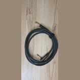 Černý instrumentální kabel s pozlacenými konektory typu jack. Na povrchu kabelu je bílý nápis GUITAR INSTRUMENT CABLE. Kabel je svázaný černým suchým…