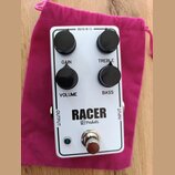 Bílý efekt pedál s nápisem RACER a logem M pedals. Na horní straně jsou čtyři černé oválné ovládací knoflíky s popisky GAIN, TREBLE, VOLUME a BASS. V…