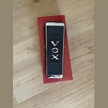 Černý kytarový wah-pedál s chromovanými okraji, umístěný na červené krabici. Uprostřed je nápis VOX a po stranách jsou texty „2 X OHMS 10W“ a „R.S.A.…