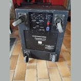 Černý aktivní subwoofer v texturovaném krytu s dvěma madly a dvěma kolečky. Na zadním panelu je ovládací sekce s konektory, potenciometry a červeným…