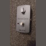 Stříbrný efekt pedál s nápisem Noise Gate a logem JOYO. Na horní části je bílý ovládací knoflík s popiskem Threshold a ve spodní části stříbrný spína…