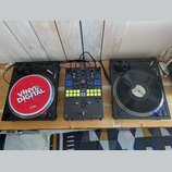 Sada DJ vybavení složená ze dvou černých gramofonů a jednoho mixážního pultu na dřevěném stole. Levý gramofon má červený slipmat s nápisy VINYL DIGIT…