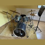 Modrá glimmer bicí souprava s chromovaným hardwarem a zlatými činely. Na basovém bubnu je bílý kožen s logem „SINCE 1970 DC DRUMS“. Na tom-tamech jso…