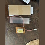 Fotografie zobrazuje elektronickou součástku s hliníkovým chladičem a přiloženou hnědou papírovou obálkou. Na oranžovém plošném spoji je bílý čtverco…