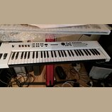 Bílá klávesová syntezátor s černými klávesami umístěná na červeném stojanu. Na levé straně je nápis YAMAHA a v pravém horním rohu označení MX61. Horn…