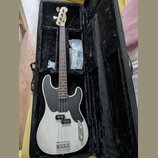 Elektrická basová kytara s bílým trupem a černým pickguardem v černém měkkém pouzdře. Na hlavě hmatníku je viditelné logo Fender. Nástroj má hnědý hm…