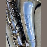 Detail stříbrného saxofonu s viditelným opotřebením povrchu a drobnými škrábanci. Na trubici jsou vyryty nápisy „H. Selmer“, „4. PLACE DANCOURT PARIS…