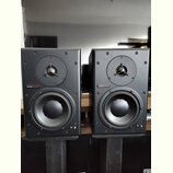Dva černé regálové reproduktory umístěné na černých stojanech. Na přední straně každého reproduktoru je nápis „dynaudio acoustics“ na červeném pozadí…