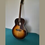 Akustická kytara s korpusem v barevném přechodu od tmavě hnědé k žluté (sunburst). Nástroj má tmavý hmatník a bílý lem kolem korpusu. Na hlavě krku j…
