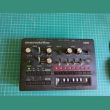 Černý kompaktní syntetizátor s nápisy monotribe ANALOGUE RIBBON STATION a KORG. Přístroj obsahuje několik otočných potenciometrů, přepínačů a řadu tl…