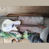 Elektrická basová kytara v krémové barvě s bílým perleťovým pickguardem. Na hlavě hnedbového krku je viditelné logo "Fender". Nástroj má čtyři struny…