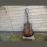 Akustická kytara s tmavě hnědým, přechodovým povrchem (sunburst) a modrým vzorovaným popruhem. Na hlavě nástroje je bílé logo Epiphone. Kytara stojí…