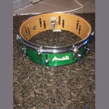 Zelený snare drum s kovovými obručemi a mechanismem na napnutí blikačů. Na korpusu je bílý nápis Amati. Nástroj vykazuje známky používání a drobné od…