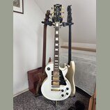 Na fotografii jsou tři elektrické kytary v držáku. V popředí je bílá kytara typu Les Paul s černým pickguardem a dvěma humbuckery, na hlavě má nápis…